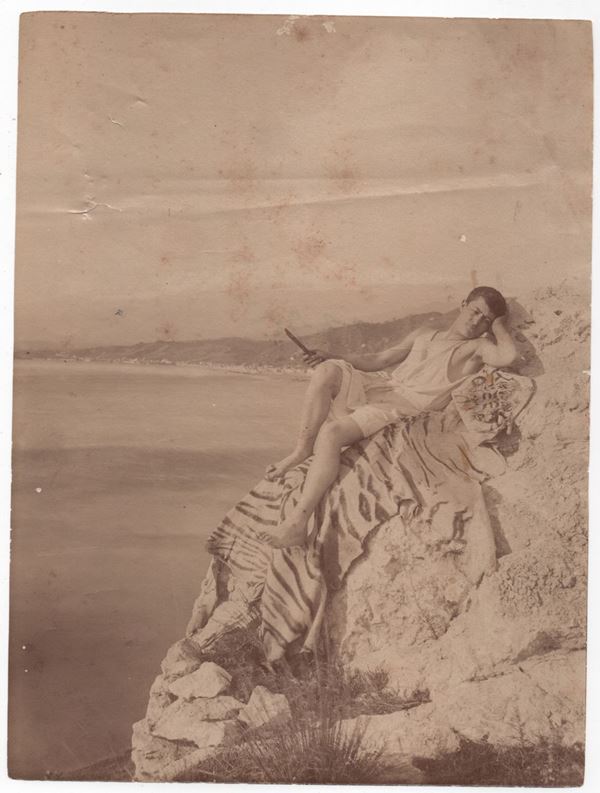 Wilhelm Von Gloeden - Ragazzo sdraiato sugli scogli con panorama di Giardini Naxos