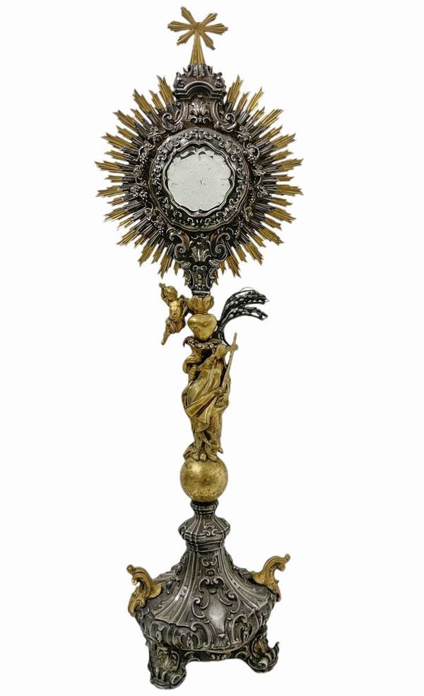 Ostensorio in  argento e argento vermeil, altezza cm 70, kg 2.352
Opera di elegante e raffinata fattura barocca in argento, eseguita a Napoli nel XVII secolo, recante i segni del saggiatore per le parti staccabili  ed il punzone con le lettere “NAP” che riguarda il periodo che va dal 1619 al 1680. La base poggia su quattro piedi a riccio con ornati vegetali a fogli d’acanto, un globo in argento dorato inciso con i simboli dell'Universo ed i segni dello zodiaco sostiene una statuina dorata raff