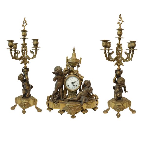 Triptych composed of a table clock in gold patinated bronze with cherubs on the sides, porcelain dial signed Imperial and a pair of candlesticks in gold patinated bronze with five lights supported by bases with putti