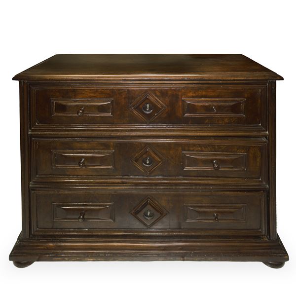 Chest of drawers with 3 drawers, rusticated and carved in a rectangle on the sides and a diamond shape in the centre with a light-coloured spout, flattened onion-shaped feet. 