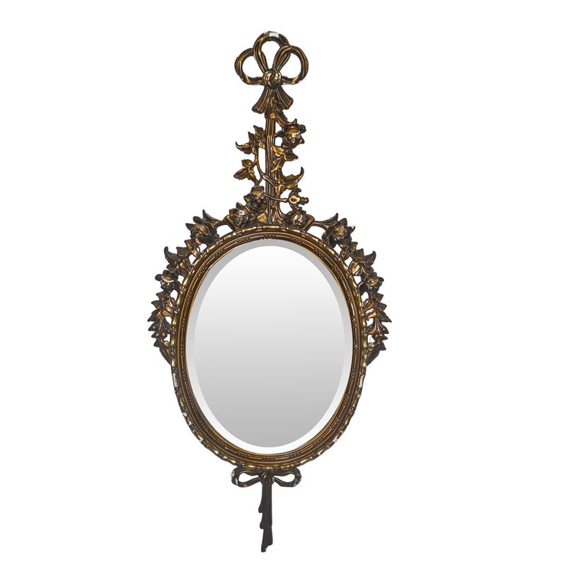 Specchiera ovale entro cornice dorata con decori floreali e fiocchi pendenti alla sommità e alla ...