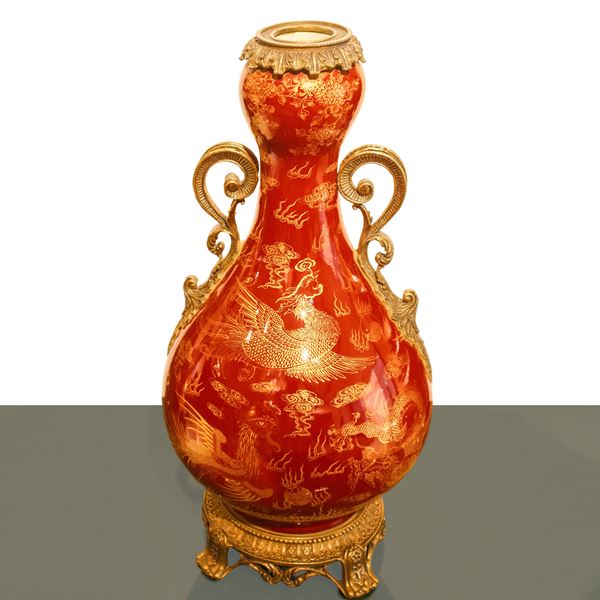 A Chinese oxblood red porcelain vase with traditional gold decorative elements and a metal base, handles and neck.