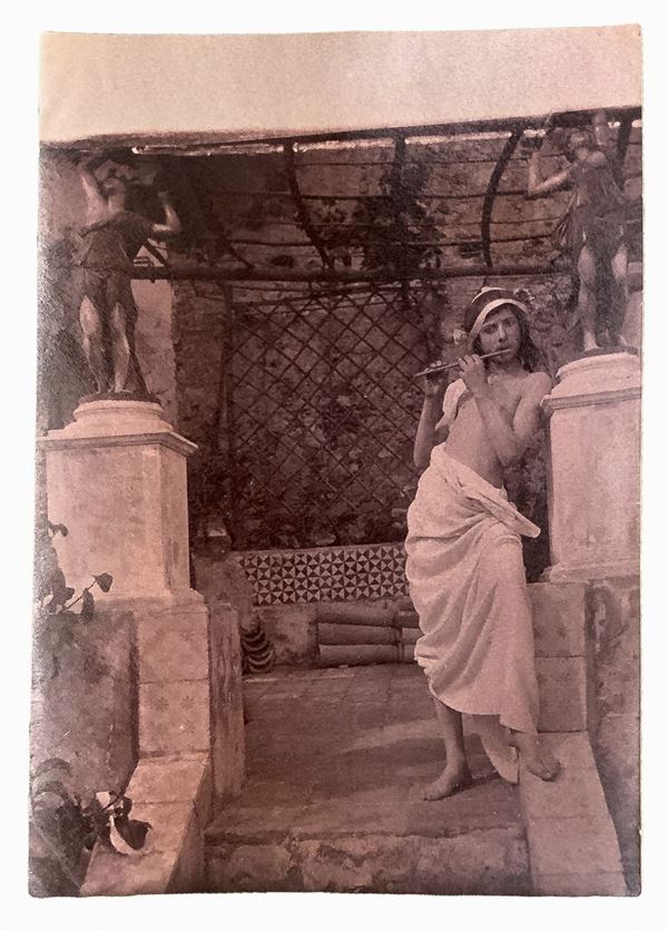 Wilhelm von Gloeden (1856-1931), foto raffigurante giovane con flauto in villa. Numerata sul retro 168 e timbrata. Cm 10,5x15

"Wilhelm Von Gloeden fu un fotografo di origine tedesca che passò parte della sua vita in Sicilia, nello specifico a Taormina, città che scelse come seconda casa. Furono i problemi di salute giovanili a portarlo nella penisola. Nello specifico la scelta di Taormina è legata all'ideale onirico della Sicilia che il fotografo sprigiona nella sua foto attraverso la scelta 